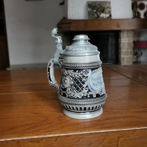 May include: A decorative beer stein with a hinged lid and handle. The stein features a detailed design with blue and silver accents. The body of the stein has a textured pattern and a decorative band around the base. The handle is ornate, and the lid is topped with a small finial.