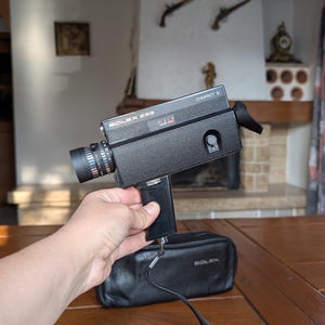 Könnte beinhalten: Schwarze Bolex 233 Compact S Filmkamera mit Objektiv und Griff. Die Kamera wird über einer schwarzen Ledertasche mit dem Bolex-Logo gehalten. Die Kamera steht auf einem Holztisch.