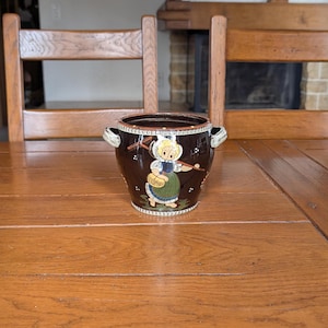 Op de afbeelding: Donkerbruine keramische pot met twee handvatten en een decoratieve illustratie van een meisje met een muts die een mand draagt. De pot heeft een witte rand en een gedetailleerd ontwerp rond de bovenkant.