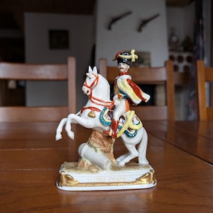 Puede incluir: Figura de porcelana de un soldado a caballo blanco encabritado. El soldado viste un uniforme colorido con capa roja y sombrero negro. La base dorada lleva la inscripción "LE PRINCE EUGENE".