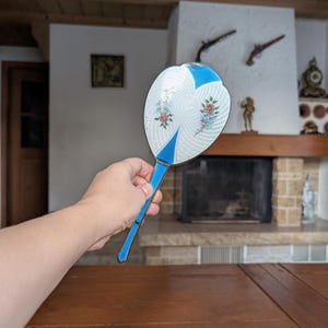May include: An antique hand fan with a white, floral design and blue accents. The fan features a silver-coloured frame and a blue handle. The fan's design includes a floral pattern with red, pink, and blue flowers. The fan is held in a person's hand.