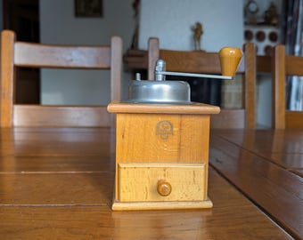 Puede incluir: Un molinillo de café de madera vintage con una parte superior de metal plateado y un mango de madera. El molinillo tiene un pequeño cajón y está sobre una mesa de madera. El fondo incluye sillas de madera y una chimenea.