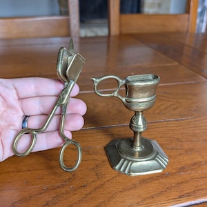 Op de afbeelding: Twee antieke messing items: een schaarachtige lonttrimmer en een kleine kandelaar. De lonttrimmer wordt in een hand gehouden. De kandelaar heeft een handvat en een zeshoekige basis. Beide items staan op een houten oppervlak.