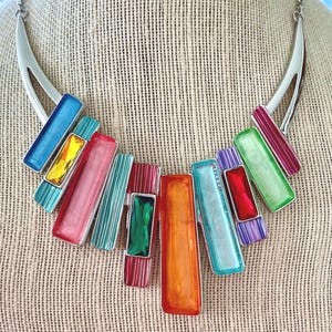 May include: A colorful statement necklace with a silver-tone metal chain and pendant. The pendant features rectangular, enameled pieces in various colors, including blue, pink, green, and orange. The necklace is displayed on a neutral-toned surface.