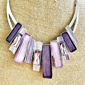 May include: A silver-toned statement necklace with a series of rectangular and geometric purple and lavender accents. The necklace features a variety of textures and shades, creating a bold and eye-catching design. The necklace is displayed on a neutral background.