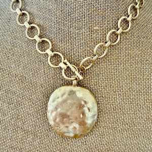 May include: A gold-toned necklace featuring a thick chain composed of circular links and a toggle clasp. A large, round, hammered-texture pendant with a metallic finish is suspended from the center of the chain, creating a bold and reflective look.