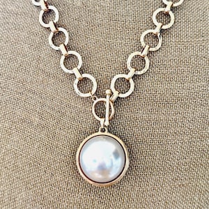 May include: A gold-toned necklace featuring a large, round, pearlescent pendant. The pendant is encased in a gold frame and hangs from a chain of interlocking circles. The necklace has a toggle clasp.