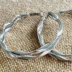 May include: A pair of silver-toned hoop earrings featuring a twisted, spiraling metal design. The earrings have a polished, reflective surface and include a standard hinged post closure mechanism at the top of each circular hoop structure.
