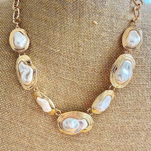 May include: A gold-tone chain necklace featuring seven irregular, white, lustrous, baroque-style faux pearl cabochons, each set within a textured, oval-shaped gold-tone metal frame. The necklace is constructed with a thick, interlocking gold-tone chain link design.