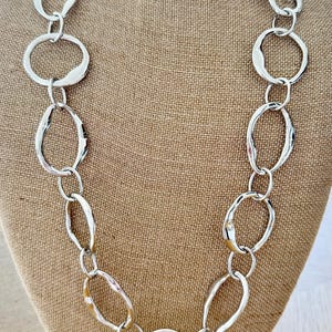 May include: A silver-toned necklace featuring a series of interconnected, irregular circular and oval links. The metal links have a smooth, reflective finish and vary in size, creating a continuous chain design that hangs in a long, draped loop.