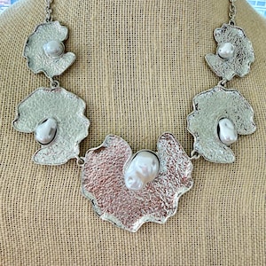 May include: A statement necklace featuring five irregular, textured silver-tone metal plates connected by a chain. The central plate has a rose-gold tone, while the others are silver. Each plate is set with a single, smooth, white baroque pearl.