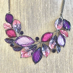May include: A necklace featuring a leaf design with a silver-tone chain. The leaves are in shades of purple, pink, and lavender, with dark purple accents. The necklace is displayed on a neutral-colored surface.