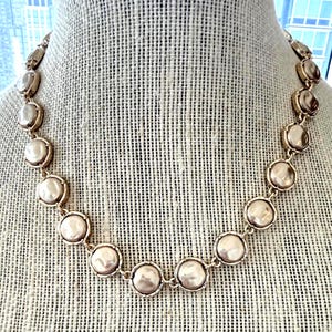 May include: A necklace featuring a series of round, lustrous, champagne-colored cabochons set in gold-toned metal frames. Each circular element is linked together to form a continuous chain, creating a uniform and repeating pattern throughout the piece.