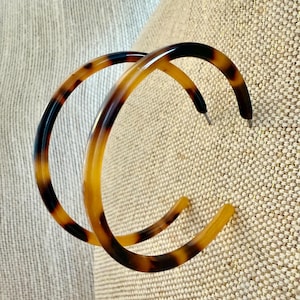 May include: A pair of large hoop earrings with a tortoiseshell pattern. The earrings feature a mix of dark brown, light brown, and amber colors. The hoops are made of a glossy material and have a simple, classic design.