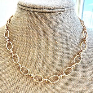 May include: A gold-toned necklace with oval links and small round beads. The necklace is displayed on a burlap-covered bust, showcasing its design and texture. The chain is made of metal and has a polished finish.