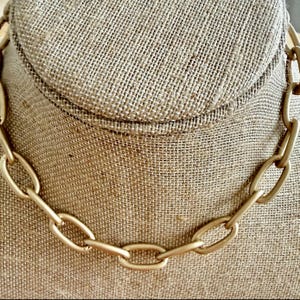May include: A gold-colored chain necklace with oval links. The necklace is displayed on a neutral-toned fabric surface, showcasing its matte finish and simple design. This piece of jewelry is suitable for everyday wear.