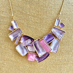 May include: A gold-toned necklace with a bib-style pendant. The pendant features a collection of geometric shapes in shades of purple and pink, outlined in gold. The necklace is displayed on a tan fabric background.