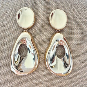 May include: A pair of gold-toned drop earrings featuring a smooth, polished finish. Each earring consists of a circular top stud connected to a larger, asymmetrical teardrop-shaped pendant with a central open cutout design.