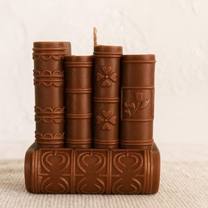 May include: A brown candle in the shape of a stack of books. The candle features a wick and is decorated with embossed designs, including shamrocks and floral patterns. The base of the candle also has an embossed design.
