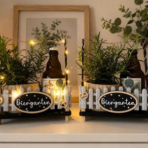 Könnte beinhalten: Zwei dekorative Biergarten-Displays mit kleinen weißen Zäunen, Holzsockeln und schwarzen Akzenten. Jedes enthält eine braune Bierflasche, eine Rosmarinpflanze und Lichterketten. Das ovale Schild trägt die Aufschrift "Biergarten".