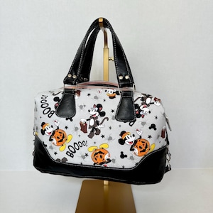Puede incluir: Bolso blanco con temática de Halloween con personajes de Mickey y Minnie Mouse, calabazas, arañas y la palabra "Boo!". El bolso tiene asas negras y una base negra.