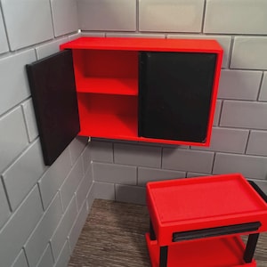Puede incluir: Un armario rojo con puerta negra y estantes interiores está montado en una pared de ladrillos grises. Debajo hay un carro rojo a juego con detalles en negro. La puerta del armario está abierta, revelando el interior rojo.