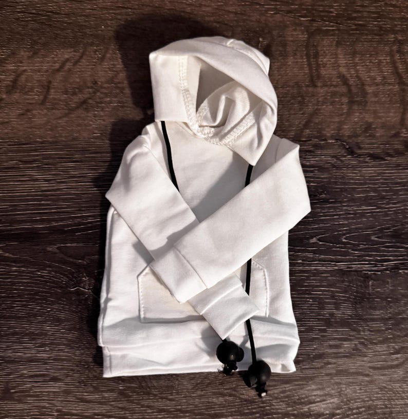 May include: A white hoodie with a drawstring and a front pocket. The hoodie has a hood and long sleeves. The drawstring is black with black ball-shaped ends. The hoodie is made of a soft, comfortable material.