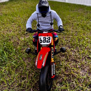 May include: A red and black mini dirt bike with the number 582 on the front. The bike has gold suspension and black tyres. The rider is wearing a grey hoodie, black gloves, and a black helmet.