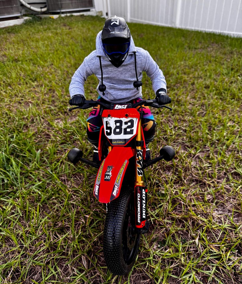 May include: A red and black mini dirt bike with the number 582 on the front. The bike has gold suspension and black tyres. The rider is wearing a grey hoodie, black gloves, and a black helmet.