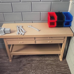 May include: A miniature workbench with a light brown surface and four drawers. Tools like wrenches and a vise are on the surface. Three colored storage bins are on the right side of the workbench. The background is a gray brick wall.