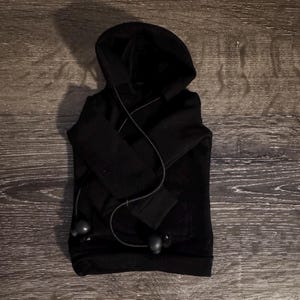 May include: A folded black hoodie with a hood and long sleeves. The hoodie has a front pocket and a drawstring. Black earbuds are draped over the hoodie.