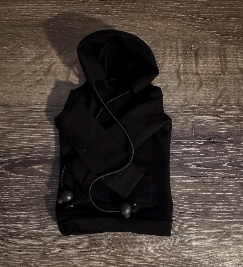 May include: A folded black hoodie with a hood and long sleeves. The hoodie has a front pocket and a drawstring. Black earbuds are draped over the hoodie.
