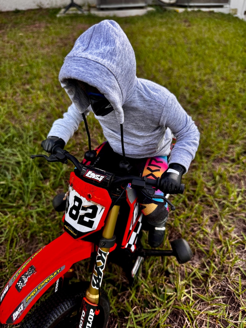 May include: A child on a red and black toy motorcycle with the number 82 on the front. The child is wearing a grey hooded sweatshirt, black gloves, and colourful trousers. The motorcycle has gold suspension forks.