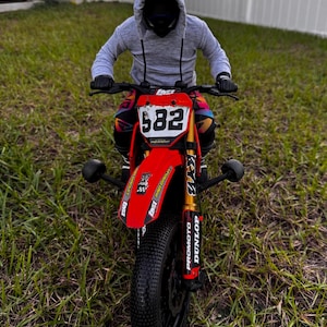 May include: A red and black motocross bike with the number 582 on the front. The bike has black tyres and gold suspension. The rider is wearing a grey hoodie and black gloves.