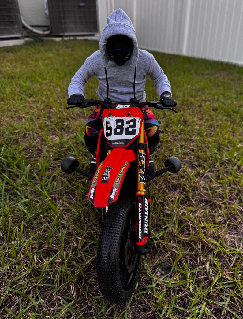 May include: A red and black motocross bike with the number 582 on the front. The bike has black tyres and gold suspension. The rider is wearing a grey hoodie and black gloves.
