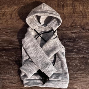 May include: A grey dog hoodie with a hood and a front pocket. The hoodie has black drawstrings and is made of a soft material. The hoodie is designed for small dogs.