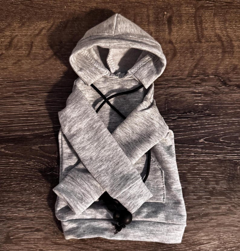 May include: A grey dog hoodie with a hood and a front pocket. The hoodie has black drawstrings and is made of a soft material. The hoodie is designed for small dogs.