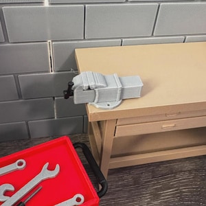 May include: A light gray miniature vise is mounted on a tan workbench. A red toolbox with silver tools sits nearby. The background features gray brick-like tiles. This image is suitable for a workshop or craft-related theme.