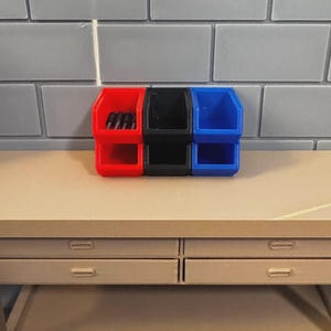 May include: A set of three small storage bins in red, black, and blue. The bins are stacked and sit on a light brown table. The background is a gray brick wall. The red bin contains small black objects.