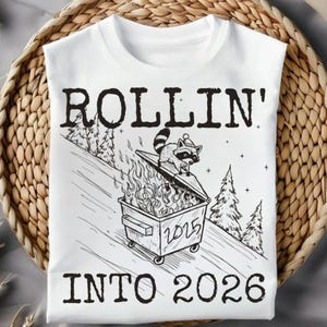 May include: White t-shirt featuring a black and white graphic of a raccoon on a burning dumpster, with the text "ROLLIN' INTO 2026". The dumpster has the year 2025 on it. The design includes pine trees.