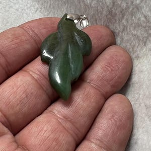 Puede incluir: Colgante de jade verde con forma de hoja. El colgante tiene una superficie pulida y una fianza plateada. El diseño de la hoja está detallado con vetas naturales. Una pieza de joyería única.