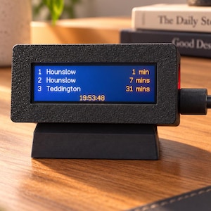May include: A black rectangular digital display device with a textured finish, featuring a blue screen showing train arrival times for Hounslow and Teddington in yellow text, with a black power cable connected to the right side and a black base.