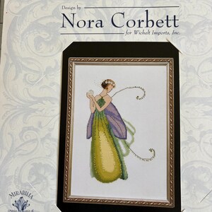 May include: A cross stitch pattern package titled Design by Nora Corbett for Wichelt Imports, Inc. featuring a Mirabilia Designs illustration of a figure in a green and purple gown with wings, holding a white sphere, framed in a gold border.
