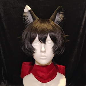 May include: A black wig with short, layered hair, styled on a mannequin head. The wig is adorned with black and white fox ears. A red scarf is tied around the neck of the mannequin, creating a striking contrast.