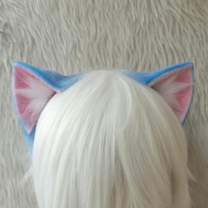 Puede incluir: Una diadema con orejas de gato azules con detalles internos rosas y blancos. Las orejas las lleva una persona con cabello blanco. Las orejas están hechas de un material suave y afelpado.