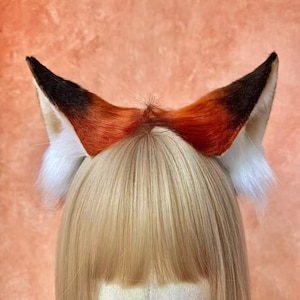 May include: A headband with fox ears. The ears are a mix of orange, brown, and white faux fur. The tips of the ears are dark brown, the base is white, and the inner part is orange. The ears are attached to a headband.