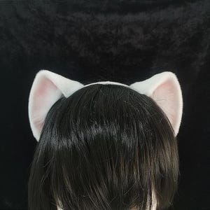 May include: White cat ear headband with pink inner ears. The ears are fluffy and stand upright, attached to a white headband. The background is a solid black color.