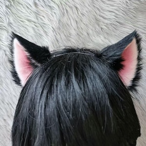 Pode incluir: Tiara de orelhas de gato pretas com orelhas internas rosa. As orelhas são feitas de um material macio e peludo e são presas a uma fina tiara preta. As orelhas são eretas e pontudas, dando um visual divertido. A tiara é usada na cabeça de uma pessoa.