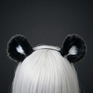 Könnte beinhalten: Ein Stirnband mit Pandaohren in Schwarz und Weiß. Die flauschigen Ohren sind schwarz mit weißen Innendetails und an einem dünnen, schwarzen Stirnband befestigt. Der Hintergrund ist dunkelgrau.