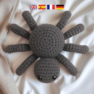 May include: A handmade, dark gray crocheted spider toy with eight legs and two small, round eyes. The spider is lying on a white surface, with a portion of a beige fabric visible in the upper left corner. A cute, soft toy.